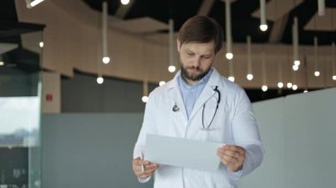 Endişeli erkek doktor elinde sağlık raporu tutarken ciddi bir ifadeyle mesafeye bakıyor..