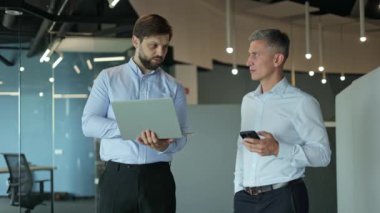 İki erkek iş adamı bir ofis ortamında dizüstü bilgisayara ve cep telefonuna bakarken işi tartışıyor.