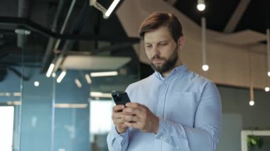 Modern bir ofis binasında telefonunu kontrol etmeye odaklanmış genç bir profesyonel. Sosyal medyada Genç Mutlu İş Adamı Mesajlaşması 'nın kapanışı. Akıllı telefon ekranına bakıyorum..