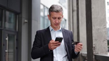 Takım elbiseli neşeli bir işadamı, açık havada duruyor, kredi kartını heyecanlı bir şekilde akıllı telefondan alışveriş yapmak için kullanıyor. Adam işlemi tamamlarken geniş bir gülümseme sergiliyor, cep telefonu ödemeleri profesyoneller.