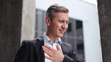 Takım elbiseli orta yaşlı bir işadamı ofisinin önünde duruyor. Göğüs ağrısının bir bölümünü yaşıyor gibi görünürken göğsünü sıkıca tutuyor. İşyeri stresi, kalp krizi veya panik atak..