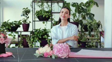 Mavi gömlekli güzel bayan çiçekçi küçük işletme sahibi arkasında pembe ve beyaz çiçeklerle dolu bir masada otururken kameraya gülümsüyor..