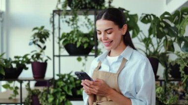 Fabrikasında çalışan genç bir kadın. Önlük takan ve telefonunu kullanırken gülümseyen bir kız. Saksı bitkileriyle dolu rafların önünde duruyor. Pencerelerden yansıyan doğal ışık..