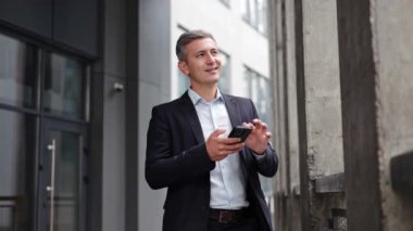 Takım elbiseli neşeli, olgun iş adamı bir şirket binasının önünde duruyor ve akıllı telefonuyla meşgul oluyor. İş iletişimi için teknolojinin kullanımını sergiliyor.