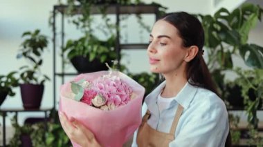 Genç ve mutlu bir kadın elinde pembe ve beyaz çiçeklerle kameraya gülümsüyor. Bitkilerle çevrili bir çiçekçide..