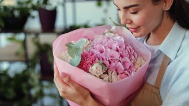 Genç bayan elinde güzel bir buket pembe çiçek tutuyor. Gülümsüyor ve çiçeklerin kokusunu alıyor. Buket pembe kağıda sarılı. Kadın mavi gömlek ve kahverengi önlük giyiyor..