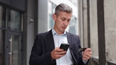 Orta yaşlı bir işadamı şirket binasının önünde duruyor ve online alışveriş için akıllı telefon kullanıyor. Odaklanıyor ve kredi kartı bilgilerini mobil bir uygulamaya aktarıyor..