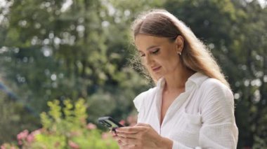 Güneşli bir günde dışarıda telefon kullanan bir kadın. Uygulamalara bakıp gülümsüyor.