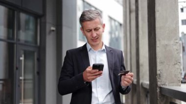 Endişeli bir işadamı, telefonuna online ödeme yapmaya çalışırken sorunlar yaşıyor. Telefonunu ve kredi kartını tutarken endişeli bir şekilde bir ofis binasının önünde duruyor..