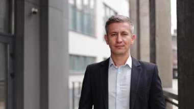 Takım elbiseli orta yaşlı bir işadamı modern bir ofis binasının önünde duruyor ve güven ve başarı yayıyor. Kamerayla göz teması kuruyor, güvenilirlik kararlılığını yansıtıyor..