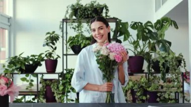 Elinde pembe ve beyaz bir buket çiçek tutarken mutlu bir çiçekçi kameraya gülümsüyor. Kadın çiçekçisinde duruyor, güzel çiçekler ve çiçeklerle çevrili.