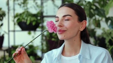 Parlak bir ofiste, çiçekli güzel bir genç kadın pembe karanfil kokusu alıyor ve gülümsüyor. O, çiçeğin kokusunun tadını çıkarmaktan mutlu. Aşk, mutluluk, bahar, tazelik ve güzellik konuları.