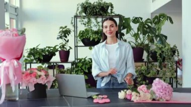 Çiçekçisinde mutlu ve arkadaş canlısı genç bir bayan çiçekçi duruyor. Etrafı bitkiler ve çiçeklerle çevrili. Kameraya sıcak bir şekilde gülümsüyor, işine güveni ve tutkusu ile bakıyor..