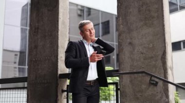Siyah takım elbiseli mutlu bir işadamı telefonunu kontrol eder, gülümser ve onaylar. Beton bir binanın merdiven boşluğunda duruyor. Bu reklam, şirket videoları, sosyal medya için mükemmel..