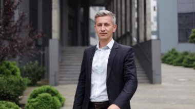 Takım elbiseli olgun bir işadamı modern bir ofis binasının önünde duruyor, gülümsüyor ve kameraya başparmak işareti yapıyor. Erkek güveni ve başarı, iş için mükemmel, finans, kurumsal temalar.
