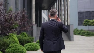 Takım elbiseli bir işadamı cep telefonuyla konuşurken kameradan uzaklaşıyor. Bir ofis binasının dışında ve asfalt bir alanda ana girişe çıkan adımlarla yürüyor..