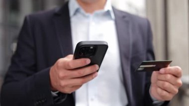 Takım elbiseli genç bir iş adamının akıllı telefonunu online alışveriş için kullanırken yakın plan fotoğrafı. Elinde bir kredi kartı var ve ayrıntıları telefonundan giriyor..