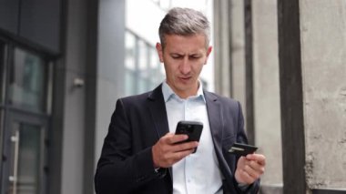 Takım elbiseli bir işadamı binanın dışında duruyor. Telefonuna kaşlarını çatmış ve elinde bir kredi kartı var. Bu da online ödemeyle ilgili bir sorun olduğunu gösteriyor..