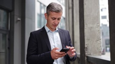 Takım elbiseli orta yaşlı bir işadamı ofis binasının önünde duruyor. Akıllı telefonuna bakarken gülümsüyor ve ekranı dinliyor, belki e-postaları okuyor ya da bir mesaj gönderiyor..