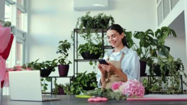 Bir çiçekçi dükkanında çiçek düzenliyor, gülümsüyor ve telefonuna bakıyor. O bir önlük giyiyor ve dükkan çok parlak ve havadar bir sürü bitki ve çiçek var..
