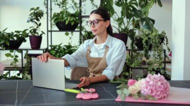 Çiçekçi olarak çalışan genç bir kadın bir sipariş için çiçek düzenler. Siparişin detaylarını kontrol etmek için masadaki bilgisayarını açıyor. Terracotta tencerelerinde odanın etrafında sayısız yeşil bitki var..