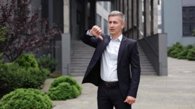İş adamı ciddi ve onaylamayan bir ifadeyle bir ofis binasının önünde duruyor. Kameraya doğru baş parmağıyla bir jest yapıyor. Onaylamadığı ya da reddedildiği açık bir mesaj veriyor..