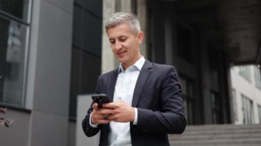 Olgun bir işadamı modern bir ofis binasının önünde duruyor ve akıllı telefon cihazını kullanmaktan mutlu. Telefonuna bakıp duruyor, sanki birini bekliyormuş ve mesaj yolluyormuş gibi..