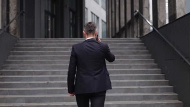 Takım elbiseli bir işadamı cep telefonuyla konuşurken modern ofis binasının dışındaki merdivenlerden yukarı çıkıyor. İş dünyası, başarı, kararlılık, odaklanma, iletişim, teknoloji veya şehir hayatı.