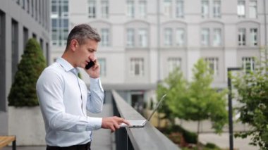 Beyaz gömlekli odaklanmış bir işadamı dışarıda duruyor, özenle dizüstü bilgisayarı üzerinde çalışıyor ve aynı anda telefonuyla bir şirket ortamını temsil ediyor..