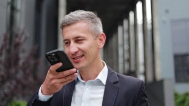 Takım elbiseli mutlu bir işadamı ofis binasının önünde duruyor. Telefonuna konuşurken gülümsüyor ve bir sesli mesaj kaydediyor. Kendinden emin ve başarılı görünüyor..