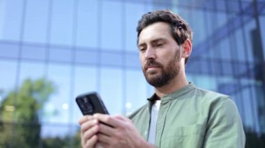 Genç bir işadamı, ofis binasının dışında telefonundaki uyarıyı kontrol ederken mutlu olur..