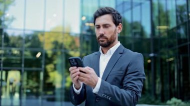 Genç bir iş adamı şehir merkezindeki modern bir ofis binasının önünde dururken akıllı telefonunu kullanmaktan mutlu olur. Şık günlük kıyafetler giymiş..