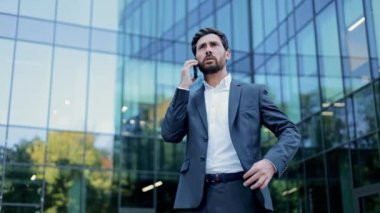 Takım elbiseli bir işadamı şirket binasının önünde duruyor ve telefonuyla konuşuyor. Mutlu ve gülümsüyor..