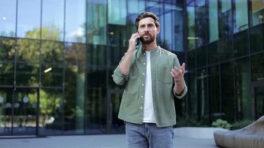 Genç bir işadamı modern bir ofis binasının önünde duruyor. Telefonla konuşuyor ve mutlu görünüyor..