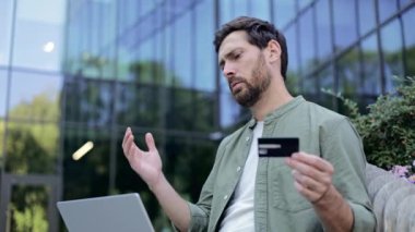 Endişeli işadamı kredi kartı tutuyor, bilgisayarında online ödemeyle boğuşuyor, stres ve hayal kırıklığı yaşıyor. İş finansmanı, çevrimiçi güvenlik ve teknoloji sorunları kavramı.