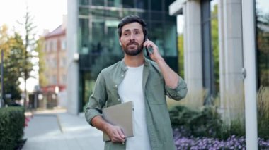 Genç bir işadamı elinde dizüstü bilgisayarla dışarıda yürüyor ve cep telefonuyla konuşuyor. Gülümsüyor ve mutlu görünüyor..