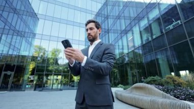 Modern bir ofis binasının önünde duran genç bir işadamı telefonundan harika haberler aldı. O, başarısını kutlamak için yumruğunu havaya kaldırır..