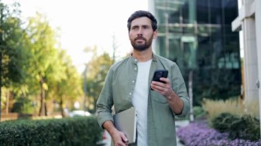 Genç bir işadamı elinde dizüstü bilgisayarla ofisin dışında yürüyor ve telefonuna gülümsüyor. Sıradan giyinmiş ve şehir manzarası çağdaş ve parlak..