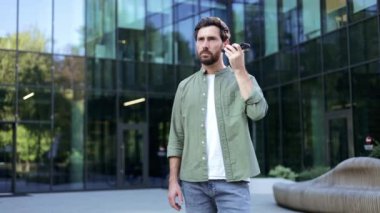 Dışarıda cep telefonuyla konuşan bir iş adamı. Sakalı var ve modern cam bir ofis binasının önünde duruyor..