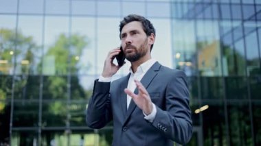 Takım elbiseli mutlu bir işadamı ofis binasının önünde telefonla konuşuyor. Gülümsüyor ve konuşurken el kol hareketi yapıyor..