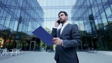 Takım elbiseli bir işadamı, modern bir ofis binasının önünde duruyor, dosyasındaki belgelere bakarken telefonuyla ciddi bir konuşma yapıyor..