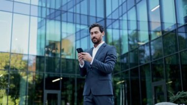 Takım elbiseli bir işadamı ofisin dışında telefonunu kullanıyor. Gülümsüyor ve sohbetten hoşlanıyor gibi görünüyor. Bu video iş, teknoloji, iletişim ve başarı temalarını betimliyor.