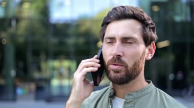 Bir iş adamı mutlu bir şekilde telefonunda sohbet ediyor. O bir ofis binasının dışında duruyor. Sakalı var ve yeşil bir gömlek giyiyor..