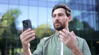 Ofis binasının dışında telefonuyla görüntülü konuşma yapan mutlu bir iş adamı. Gülümsüyor ve el sallıyor..