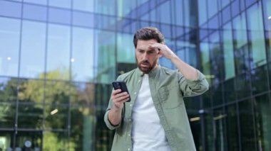 Genç bir işadamı, akıllı telefonuyla ilgili haberler aldı. Bir ofis binasının önünde duruyor ve hayal kırıklığını açıkça gösteriyor..