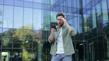 Bir şirket binasının önünde duran genç bir profesyonel telefonundaki mesajı okurken şaşkın ve endişeli görünüyor..