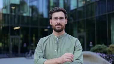 Gözlüklü ve sakallı genç bir işadamı modern bir ofis binasının önünde kollarını kavuşturarak kameraya gülümsüyor. Güven ve başarı saçar..
