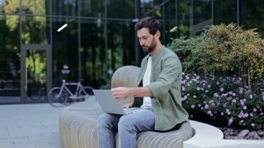 Ofis binasının dışındaki bankta oturan bir iş adamı dizüstü bilgisayarıyla çalışıyor. Günlük kıyafetler giyiyor ve sakalı var. Adam çalışırken gülümsüyor..