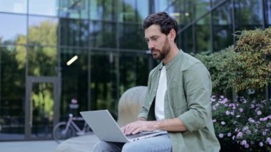 Genç bir iş adamı laptopuyla ofisinin önünde çalışıyor. Bilgisayarda çalışırken gülümsüyor, başarılı bir iş anlaşması ya da başarı öneriyor..