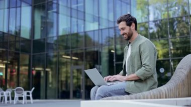 Genç bir işadamı güneşli bir günde ofis binasının dışındaki bankta oturur ve dizüstü bilgisayarıyla çalışırken gülümser. O mutlu ve üretken..